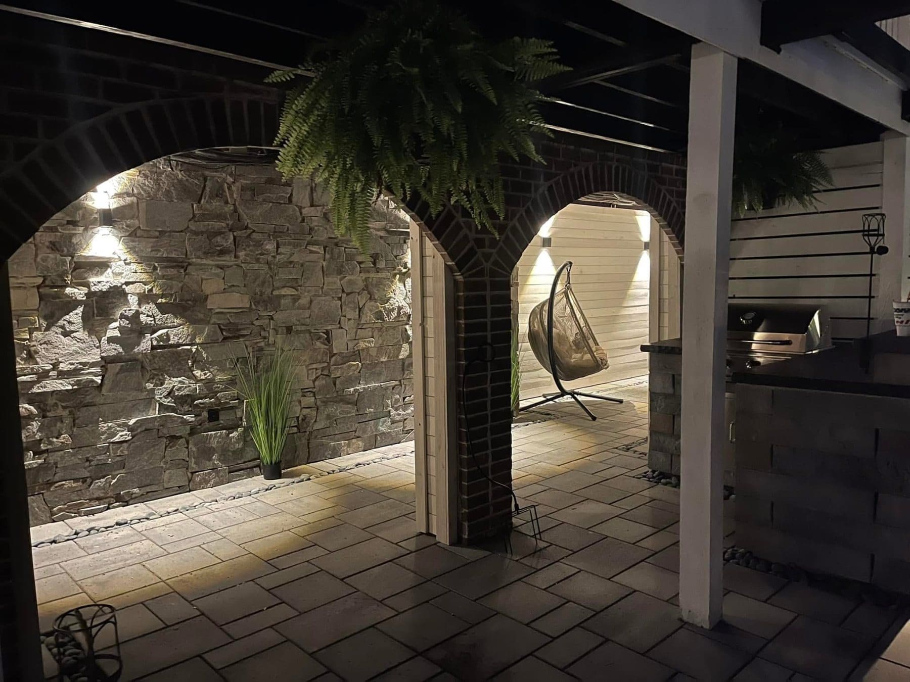Covered patio with stone archway, hanging fern, swing chair, and ambient lighting at night