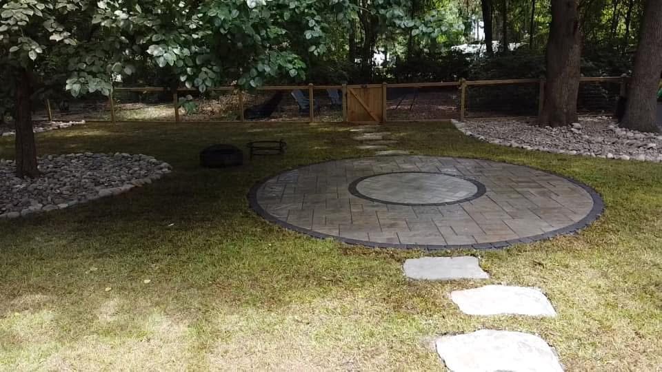Circular paver patio with fire ring surrounded by lawn and wooden fence in backyard