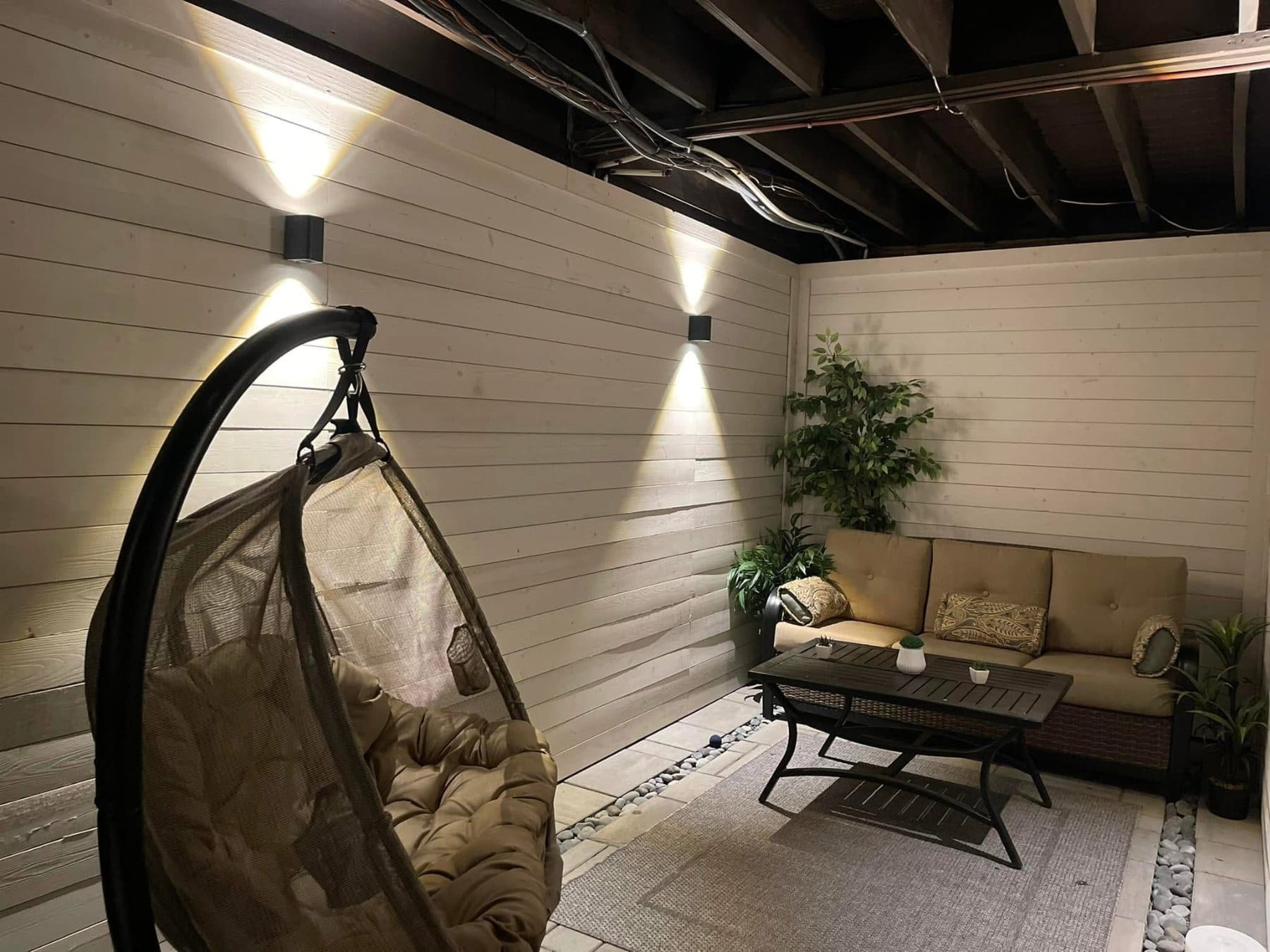 Modern patio space with hanging chair, couch, coffee table, plants, and wall-mounted lights