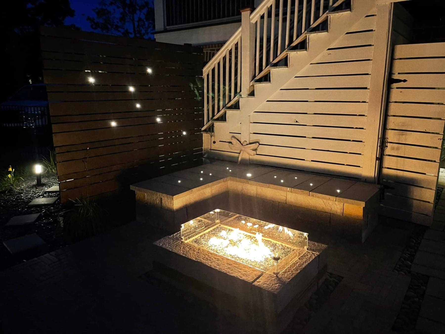 Illuminated outdoor patio at night featuring a sunken fire pit, built-in seating, and wooden fencing