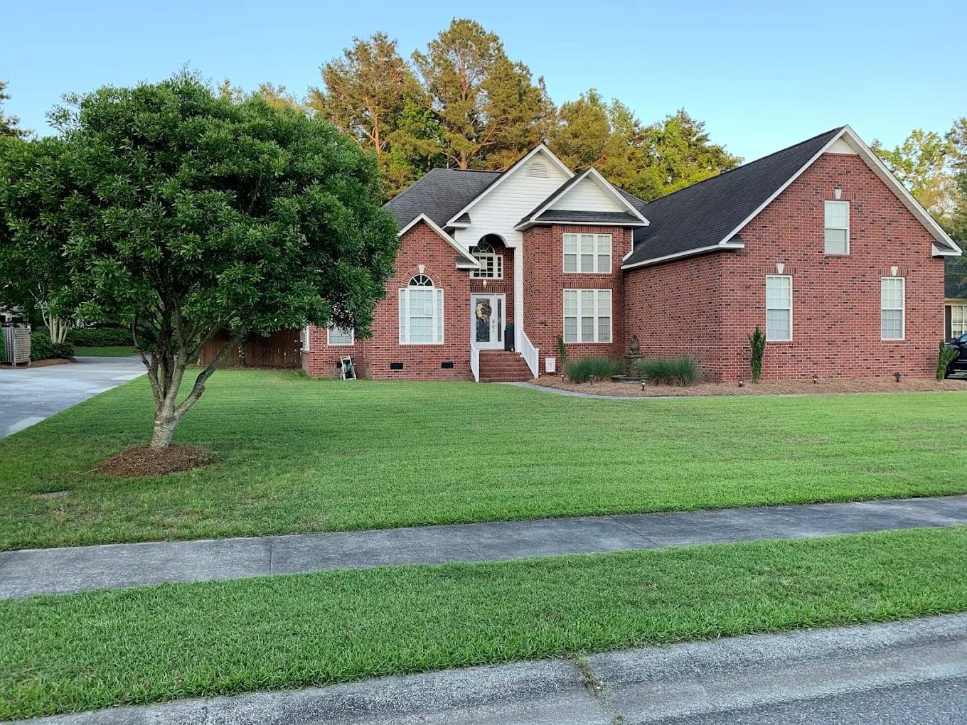 Red brick single-story house with white trim, manicured lawn, driveway, and mature trees
