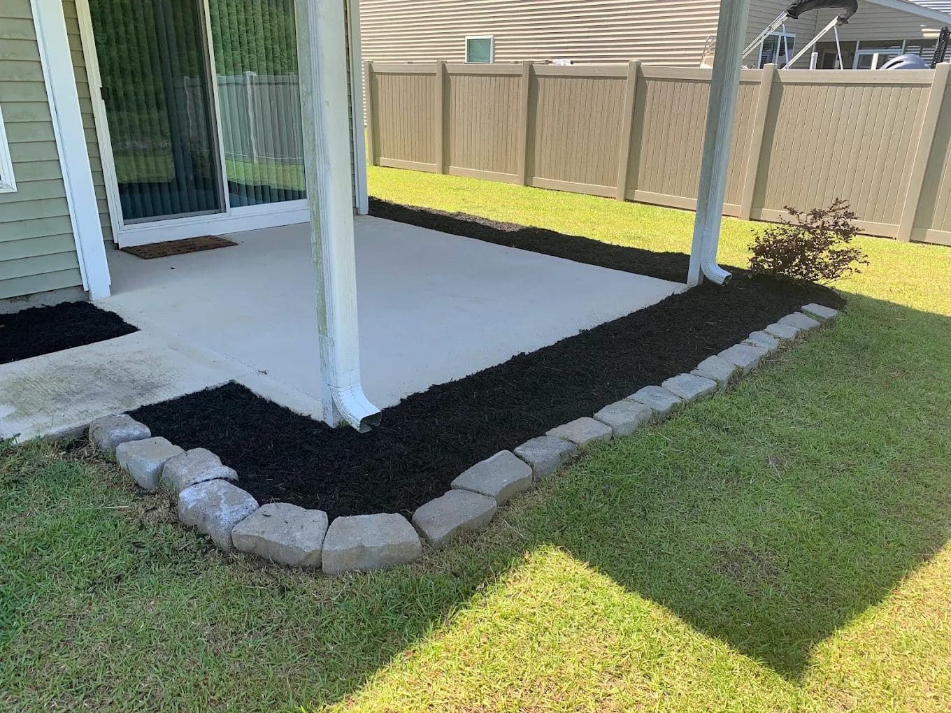 Backyard patio with concrete slab, mulch garden beds, and decorative stone edging along lawn