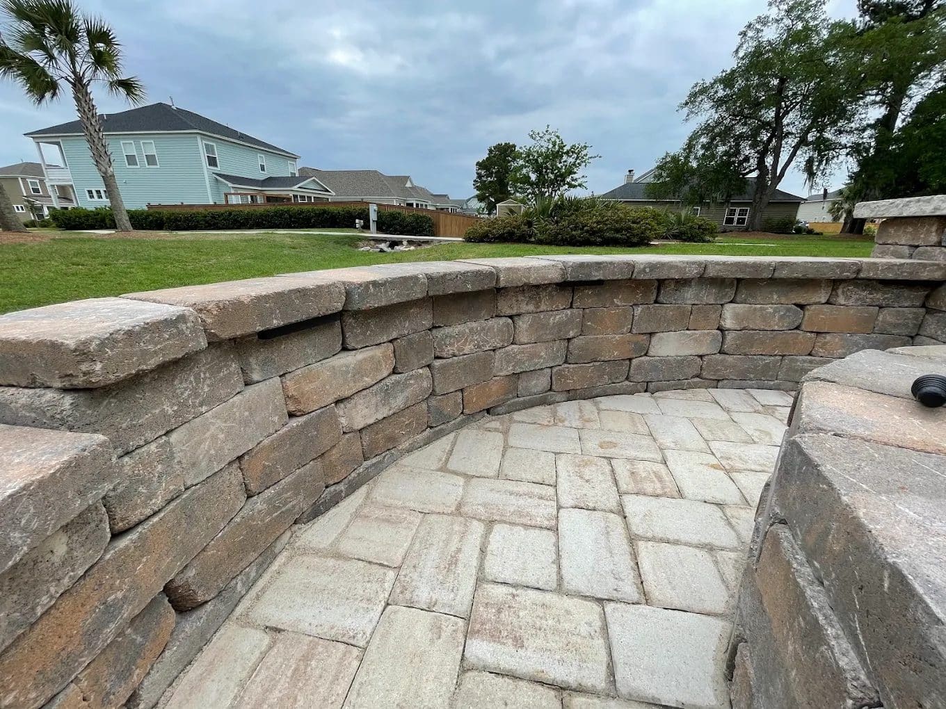 Curved stone retaining wall with paver patio overlooking residential neighborhood with houses and green lawn